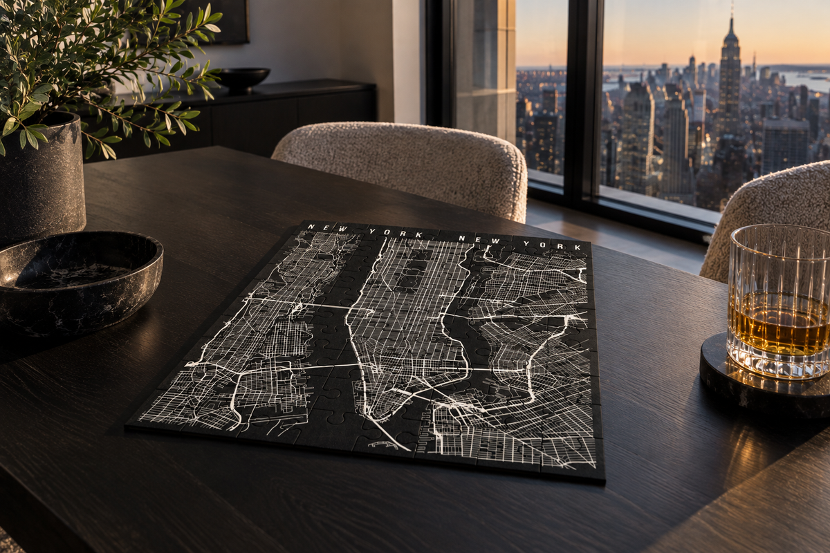 A black rectangular NYC metal jigsaw puzzle with white street-line artwork is displayed on a dark wooden table in a modern room, with the New York City skyline visible through large windows behind it. A glass of amber drink and chairs are also present in the scene.