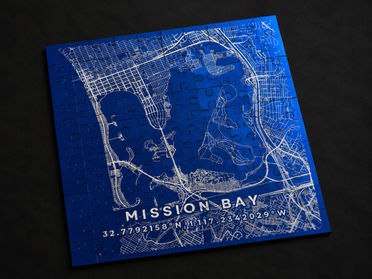 Blue anodized aluminum Mission Bay map jigsaw puzzle with white street lines and coordinates, lying flat at an angle on a dark background.