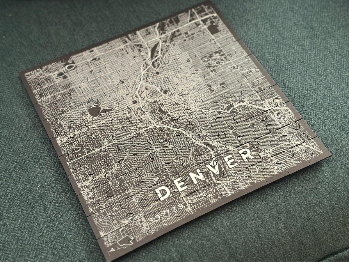 A square jigsaw puzzle featuring a vintage-style street map of Denver, Colorado, with the city name etched in white at the bottom, resting on a textured dark green fabric surface.