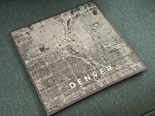 A square jigsaw puzzle featuring a vintage-style street map of Denver, Colorado, with the city name etched in white at the bottom, resting on a textured dark green fabric surface.